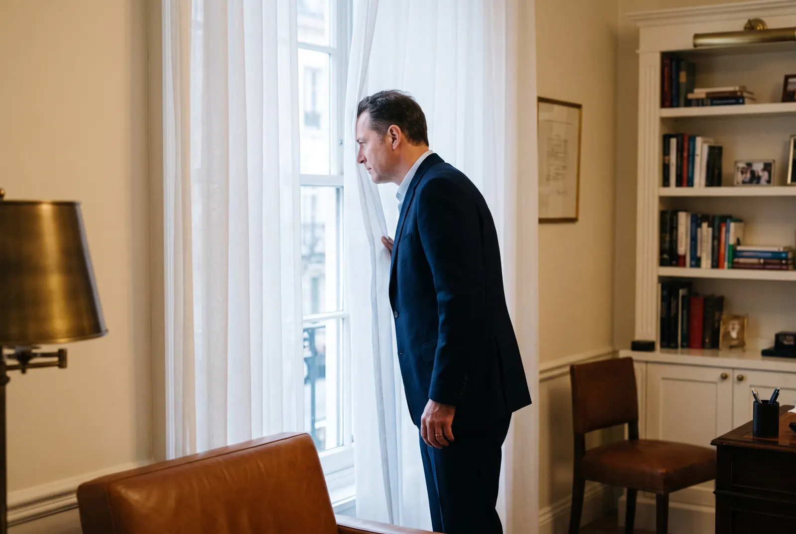 A man in a navy suit stands behind sheer curtains looking out a tall window